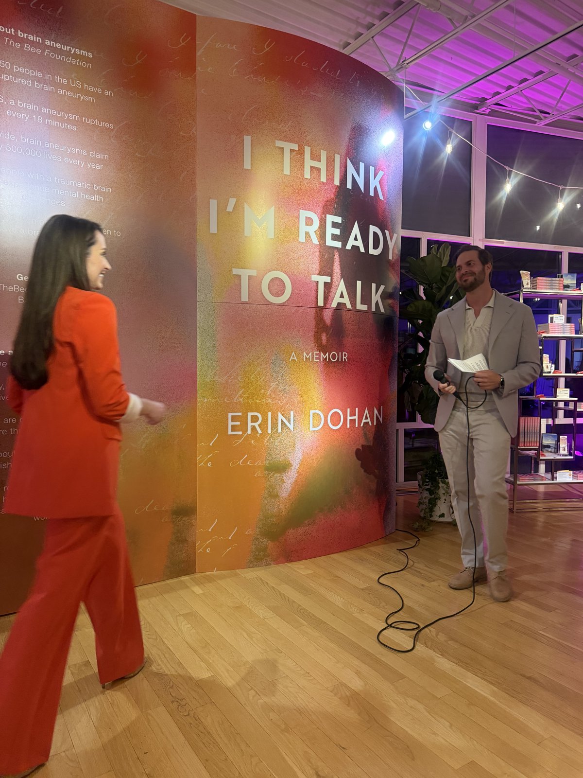 Patrick at the microphone with his speech on paper, Erin walking up beside the book wall